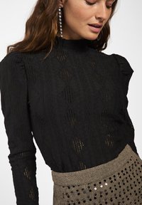 Vrouw in een zwarte, gestructureerde lange mouwtop met diamantpatronen en een beige high-waisted rok met zwarte pailletten, die iets naar beneden kijkt.