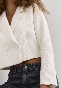 Kürzer geschnittene, doppelt lose Jacke aus strukturiertem weißen Stoff, mit großen Revers und weiten Ärmeln, kombiniert mit hoch taillierten dunklen Jeans.