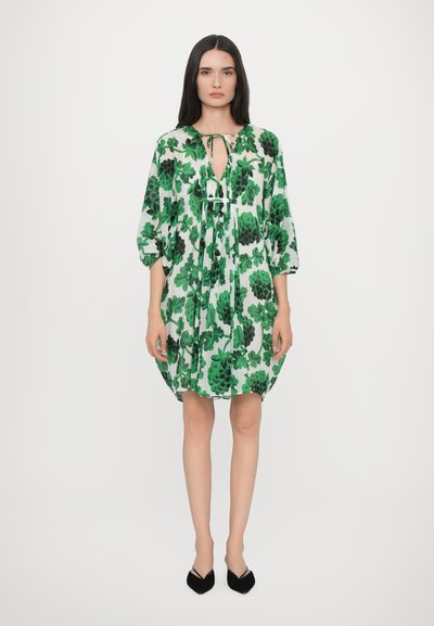 Robe florale avec des motifs de feuilles vertes sur une base blanche, dotée d'un décolleté en forme de clef et de manches amples, assortie à des talons noirs.