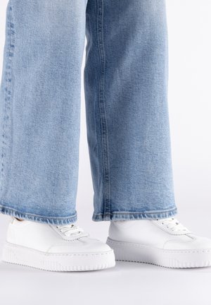 Person, die weit geschnittene hellblaue Jeans und weiße Schnürplattform-Sneaker trägt, steht auf einer weißen Oberfläche.