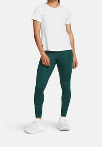 Kobieta w białej koszulce z krótkim rękawem, ciemnozielonych legginsach i białych butach sportowych stoi na jednolitym tle.