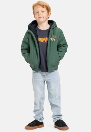 Jongentje met rood haar, gekleed in een groene jas, een donker grafisch T-shirt, lichtblauwe spijkerbroek en zwarte sneakers, staande met zijn handen in zijn zakken.