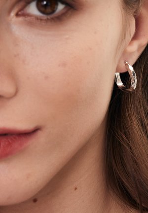 Srebrna kolczyk w formie obręczy z teksturowanym wzorem, posiadający gładkie wykończenie i mały detal akcentujący, noszony na płatku ucha modelki.