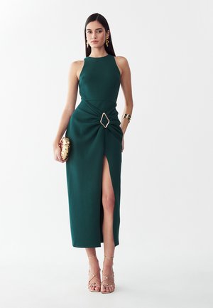 Vrouw draagt een mouwloze donkergroene jurk met een split aan de voorkant, gouden gesp, gouden clutch, gouden oorbellen, armband en sandalen met bandjes.