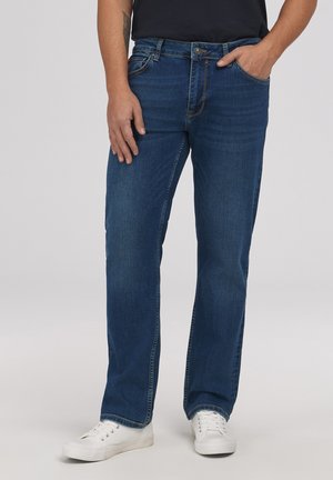 Mężczyzna w niebieskich dżinsach o prostych nogawkach i białych trampkach, z jedną ręką w kieszeni, a drugą luźno opartą przy boku.