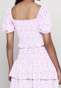 Una persona indossa un top floreale lilac aderente con maniche a sbuffo, abbinato a una gonna a strati coordinata. Lo sfondo è grigio uniforme.