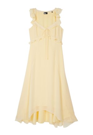Jeune femme portant une robe asymétrique sans manches jaune pâle avec des volants et des sandales compensées blanches, debout devant un fond uni.