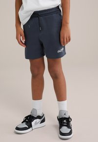 Námořnické modré šortky s elastickým pasem a černou šňůrkou; obsahují bílé logo "SALTY". S kombinací černých a šedých sportovních bot.