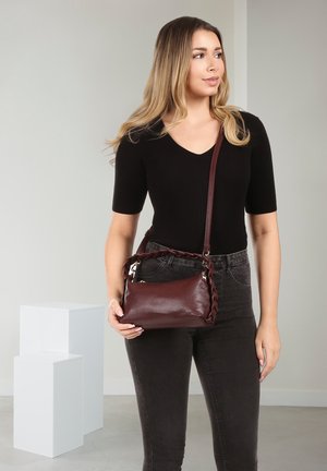 Woman in black top and jeans holding a brown leather crossbody bag with braided detailing, standing near white display blocks.