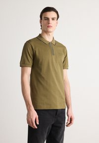 Olive zaļš polo krekls, kas izgatavots no mīksta kokvilnas, ar rāvējslēdzēju, īsām piedurknēm un maigi melnām dekoratīvajām līnijām ap apkakli.