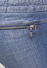 Gros plan sur un tissu en denim bleu avec une fermeture éclair métallique horizontale et une petite languette portant l'étiquette "GUESS" cousue dans la couture.