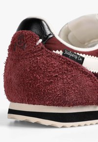 Zapatilla burdeos con material texturizado y suave, tacón de cuero negro y detalles en blanco. Presenta un logo y una suela acanalada de color crema.