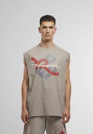 Chemise sans manches beige avec un grand design graphique rouge et gris et le texte "Rocawear" en blanc. Fabriquée en tissu doux et léger.