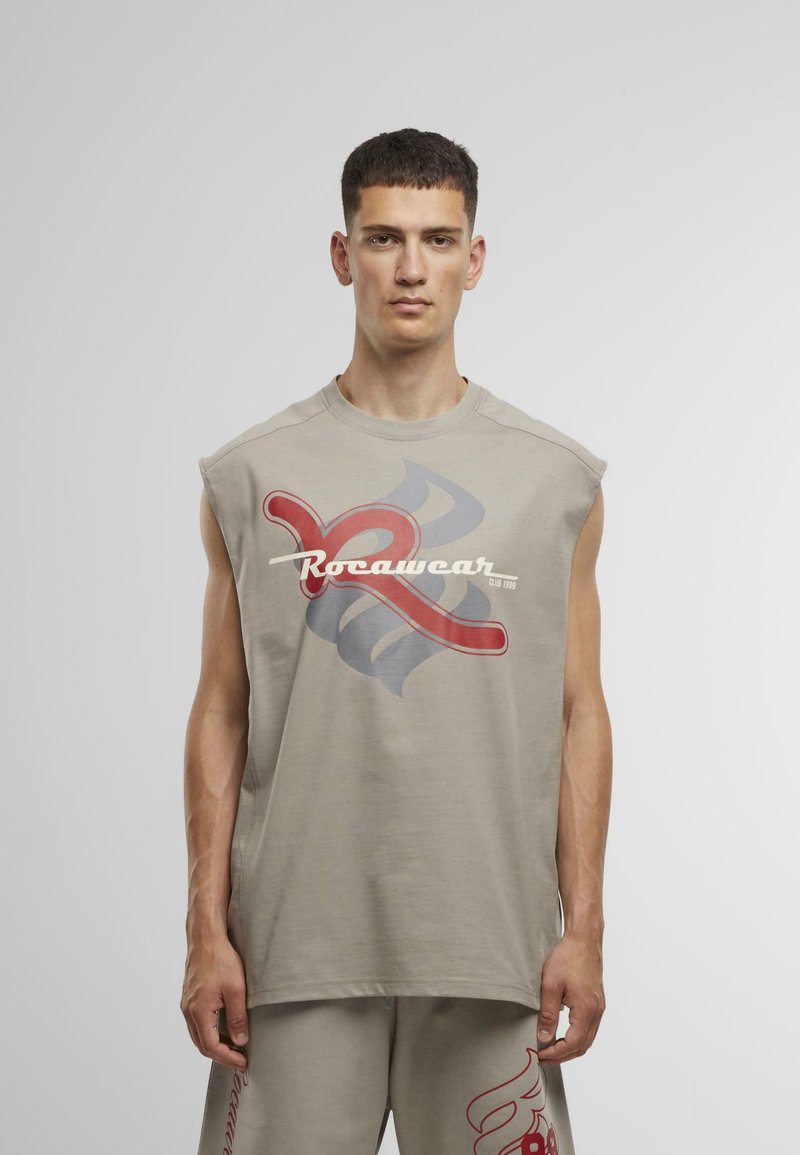 béžová bezrukávová košieľka s veľkým červeným a šedým grafickým dizajnom a bielym textom "Rocawear". Vyrobená z mäkkej, ľahkej tkaniny.
