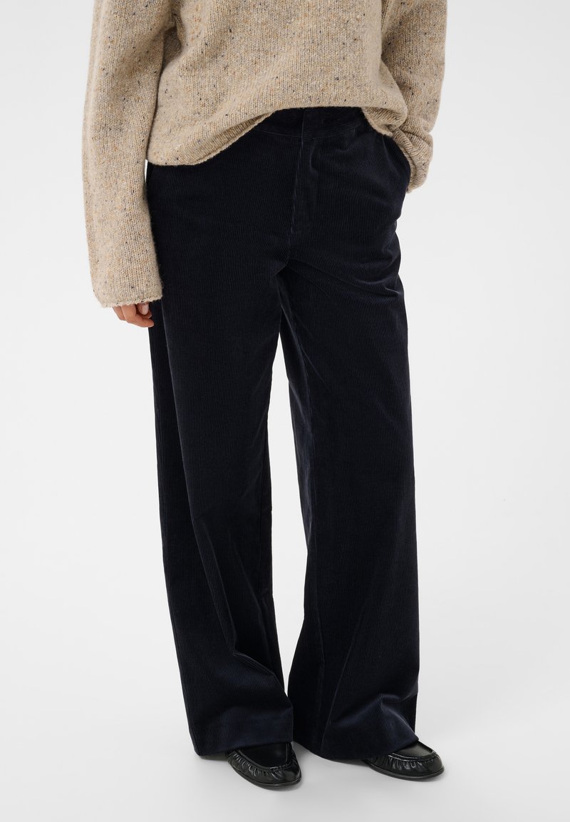 Personne portant un pantalon large en velours côtelé noir, un pull en tricot beige et des chaussures plates noires, debout avec une main dans la poche.