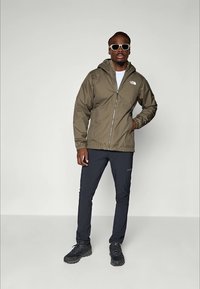 Homme portant une veste à capuche olive, une chemise blanche, un pantalon noir, des chaussures de randonnée noires et des lunettes de soleil blanches, se tenant devant un fond uni.