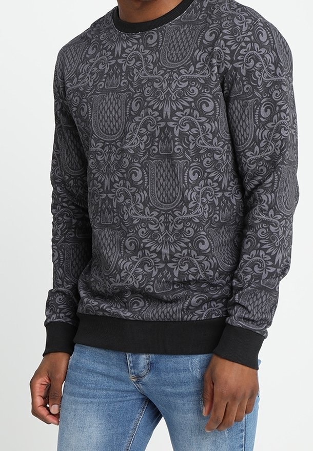 Homme portant un pull sombre à manches longues avec des motifs floraux et géométriques complexes et un jean bleu clair, debout devant un fond blanc.