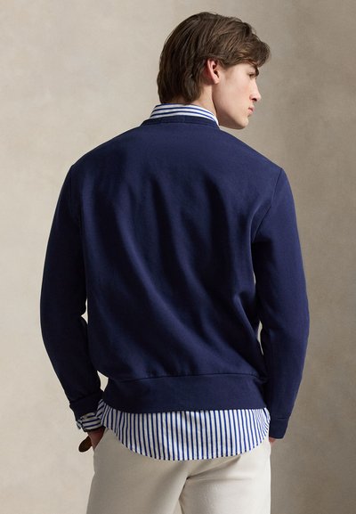 Homme de profil portant un pull bleu marine par-dessus une chemise rayée bleue et blanche avec un pantalon blanc sur un fond neutre.