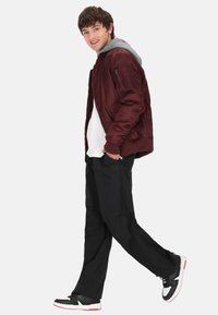 Bordeaux bomberjakke med grå hætte, hvid t-shirt, løse sorte bukser og sorte sneakers med røde detaljer. Afslappet, løs pasform.