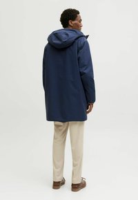 Marineblauer wasserdichter Mantel mit Kapuze, geradem Schnitt und Rückenschlitzdetails, kombiniert mit beigen Hosen und braunen Schuhen.