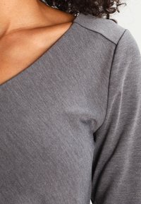 Maglietta grigia a maniche lunghe con scollo ampio, texture liscia e un delicato motivo a coste, che mette in evidenza una vestibilità rilassata sulle spalle.
