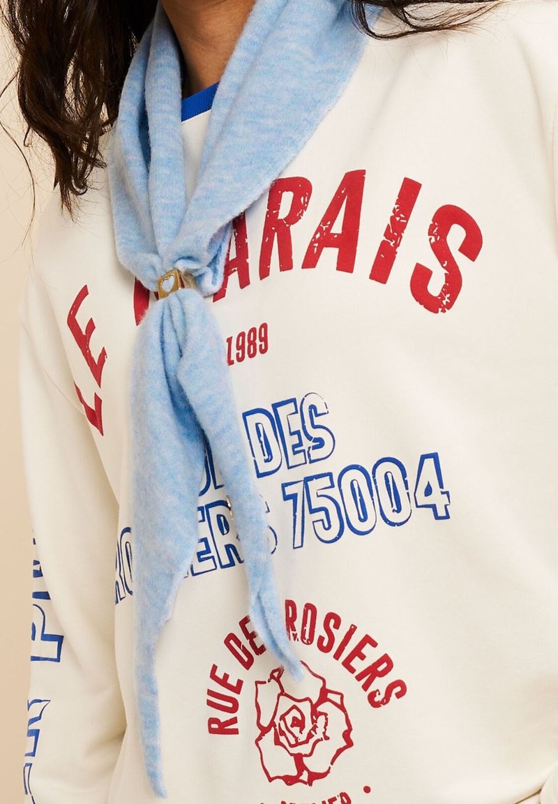 Person trägt ein weißes Sweatshirt mit rotem und blauem Paris-Motiv-Text und einen hellblauen Schal, der mit einer Anstecknadel befestigt ist.