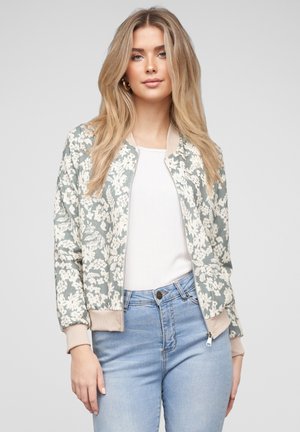 Jonge vrouw met lang blond haar draagt een bomberjack met bloemenpatroon, een wit topje en lichtblauwe high-waisted spijkerbroek tegen een grijze achtergrond.