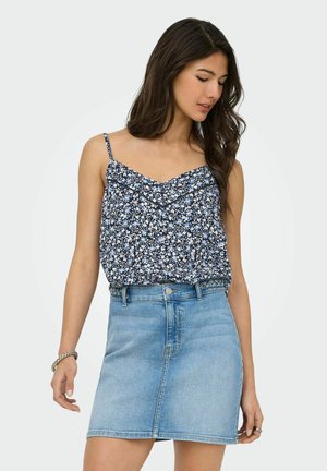 Vrouw met lang donker haar in een marineblauwe bloemen mouwloze top en een lichtblauwe spijkerrok, staand en lichtjes naar beneden en opzij kijkend.