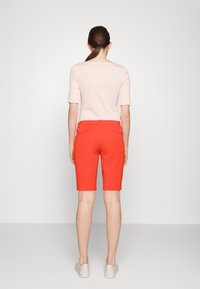 Kvinna med brunt hår i hästsvans som bär en ljusrosa kortärmad topp, ljusröda knälånga shorts och vita sneakers, står vänd bort.