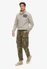 Grå fleecepullover med høj krave og marineblå logo. Camouflage cargo bukser i grønne nuancer med flere lommer. Lysebrune høj-top sneakers.