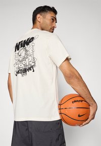 Cremefarvet kortærmet t-shirt med et sort grafisk print på ryggen, der viser et basketball-design. Modellen holder en basketball.