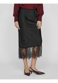 Jupe crayon noire en satin avec un ourlet en dentelle noire transparente présentant des motifs floraux complexes. Portée avec des talons pointus.