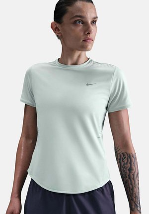 Femme aux cheveux tirés en arrière portant un t-shirt à manches courtes gris clair de Nike, montrant un tatouage détaillé sur son avant-bras gauche.