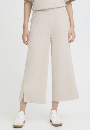 Mujer con pantalones beige de pierna ancha y corte cropped con aberturas laterales, combinados con zapatillas blancas, posando frente a un fondo blanco.