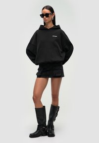 Czarna, oversize'owa bluza z kapturem z kieszenią z przodu, zestawiona z czarną mini spódniczką dżinsową z kieszeniami cargo. Czarne kozaki za kolano z klamrami.