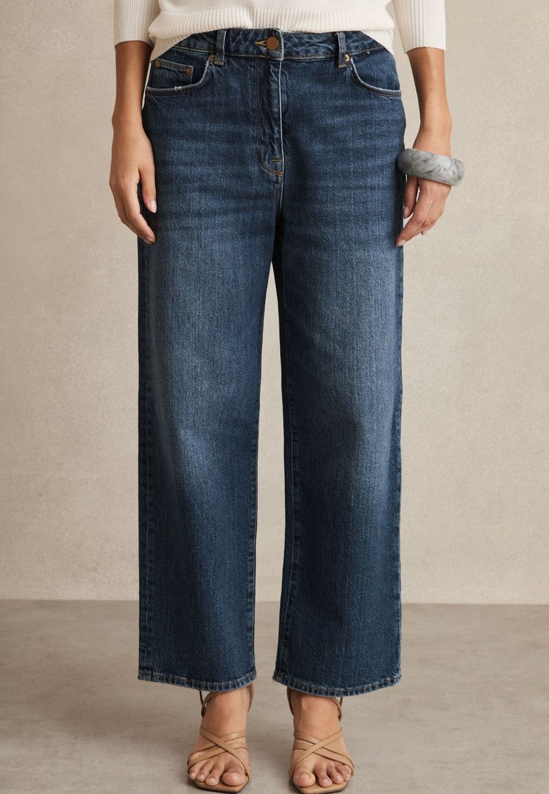 Vooraanzicht van een persoon die hooggetailleerde, rechte blauwe jeans, beige sandalen met bandjes, een witte trui en een chunky grijze armband draagt.