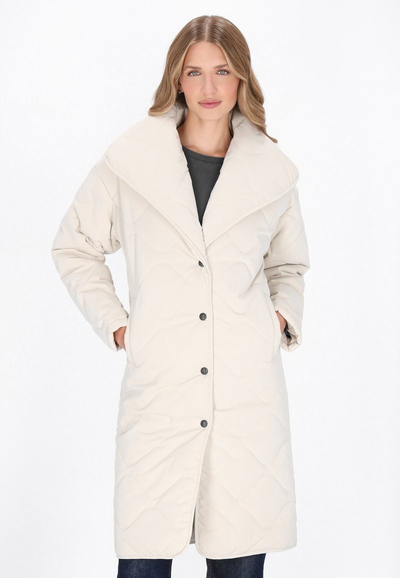 Hosszú, steppelt kabát világos bézs színben, nagy gallérral, elülső gombokkal és két oldalsó zsebből áll. Puha textúrájú, sima felülettel.