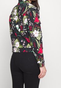 Giacca a stampa floreale con sfondo nero, fiori rossi e bianchi vivaci, foglie verdi e maniche lunghe con un design aderente.