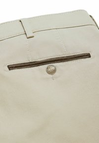 Close-up van beige broeken met een achterzak met een knoopsluiting en een riemlus boven de zak.