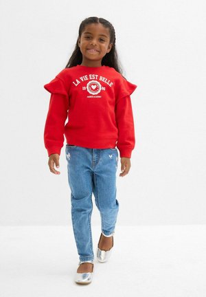 Felpa rossa con maniche arricciate e testo bianco, abbinata a jeans blu chiaro con accenti ricamati e scarpe argento lucido.