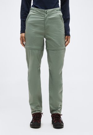 Pohľad spredu na osobu v oliva zelených konvertibilných turistických nohaviciach a čiernych turistických topánkach, nohavice sú nad kolenami zipsom odopnuté a vytvárajú šortky.