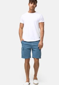 T-shirt en coton blanc et shorts cargo bleus avec poches latérales. Les chaussures sont des baskets blanches à enfiler. Tenue d'été simple et décontractée.