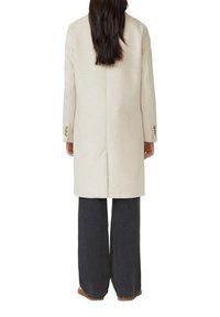 Manteau en laine couleur crème au design ajusté, avec des manches longues et des accents de boutons, associé à un pantalon large noir et des chaussures marron.
