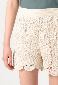 Lace shorts i en cremet farve med blomsterprint, der har en tekstureret talje og bølget kant, vist på en models hånd.