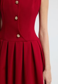 Vestido rojo sin mangas con un corpiño ajustado, botones de textura dorada a lo largo del frente y una falda plisada que se muestra desde el hombro hasta la cintura.