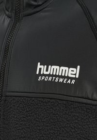 Close-up van zwarte sportkledingstof met doorgestikte en fleece texturen, met wit "hummel SPORTSWEAR" logo en ritsdetail.