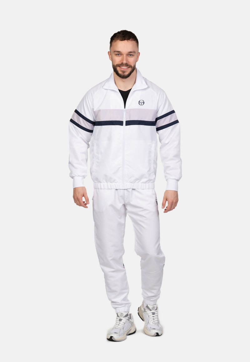 Witte trainingspak met een ritsjack dat navy en grijze strepen heeft. De broek heeft elastische boorden. Model draagt witte sneakers.