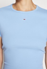 Zbliżenie osoby noszącej jasnoniebieską prążkowaną koszulkę z krótkim rękawem, na której w centrum, tuż przy kołnierzu, znajduje się małe czerwono-biało-granatowe logo.