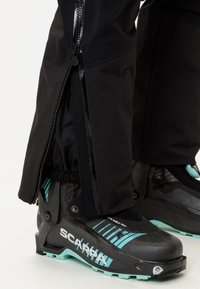 Personne portant un pantalon noir avec une fermeture éclair latérale sur des chaussures de ski Scarpa noires et turquoises.