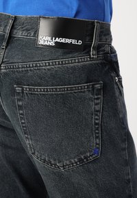 Crne traperice od denima s kožnom oznakom s markom "KARL LAGERFELD JEANS" i malim plavim detaljem blizu stražnjeg džepa.
